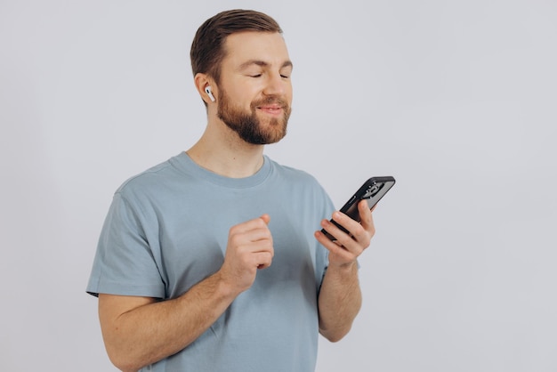 Homem barbudo feliz dançando com fones de ouvido ouvindo música em fones de ouvido segurando o celular e sorrindo satisfeito em pé sobre fundo branco