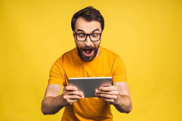 Homem barbudo feliz espantado usando tablet digital, parecendo chocado com as notícias da mídia social, consumidor de comprador homem espantado surpreso com a vitória online isolada sobre fundo amarelo.