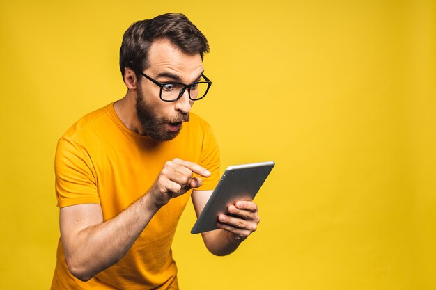 Homem barbudo feliz espantado usando tablet digital, parecendo chocado com as notícias da mídia social, consumidor de comprador homem espantado surpreso com a vitória online isolada sobre fundo amarelo.