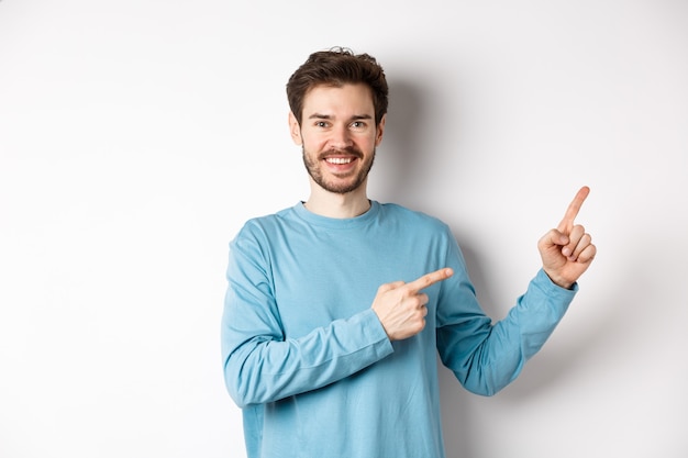 Homem barbudo feliz mostrando o anúncio, apontando os dedos para a direita e sorrindo, convidando para clicar no link, em pé sobre um fundo branco.