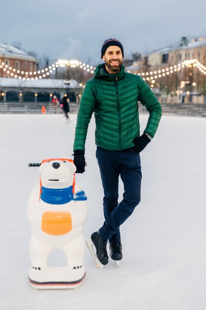 Homem barbudo feliz usa jaqueta verde e chapéu, fica perto de ajuda de skate como tentar obter habilidades de patinação