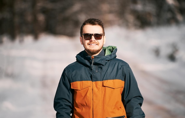 Homem barbudo feliz usando óculos escuros em clima ensolarado de neve Conceito de passar um tempo ativo caminhando após uma nevasca O cara sorrindo e vestindo uma jaqueta de inverno