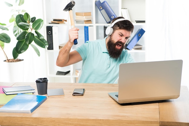 Homem barbudo fones de ouvido escritório balanço martelo no computador Conexão lenta com a internet Software desatualizado Atraso do computador Razões para o atraso do computador Como corrigir o sistema de atraso lento Odeie a rotina do escritório