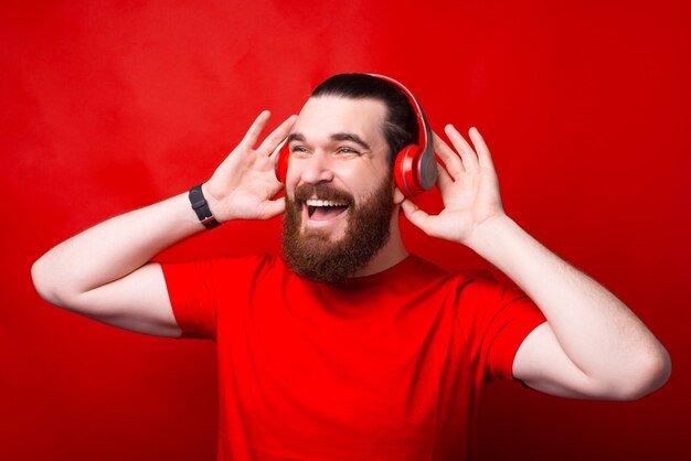 Homem barbudo hippie alegre ouvindo música em fones de ouvido na parede vermelha