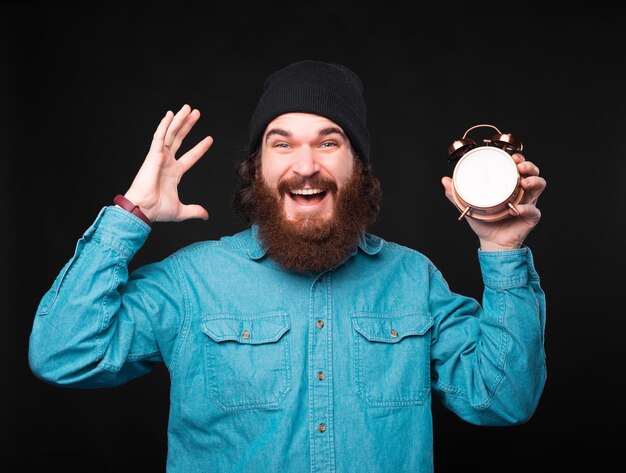 Homem barbudo hippie espantado segurando um despertador sobre um fundo escuro