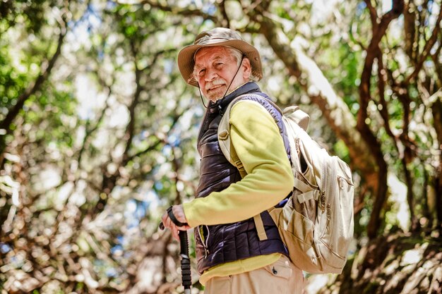 Homem barbudo idoso alegre com mochila e chapéu caminhando na floresta desfrutando de um estilo de vida saudável