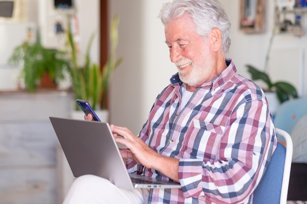 Homem barbudo idoso sentado em casa segurando telefone celular Alegre e sorridente belo homem sênior desfrutando de tecnologia e social