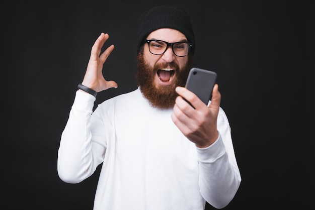 Homem barbudo irritado, gritando com smartphone em pé sobre fundo escuro