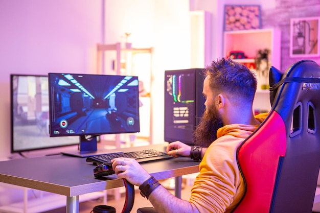 Homem barbudo jogando videogame online em seu pc e conversando com outros jogadores. Neons coloridos na sala.