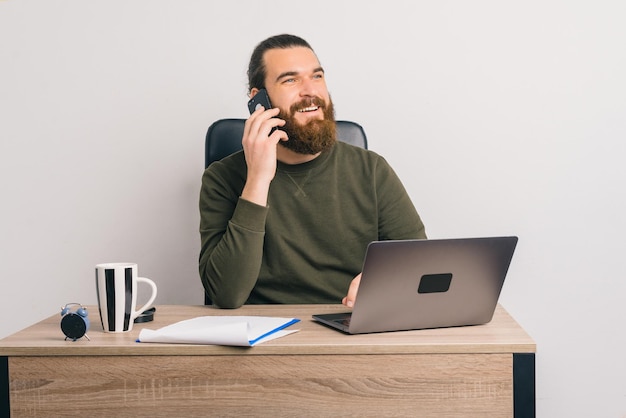 Homem barbudo jovem amigável está falando ao telefone no escritório.