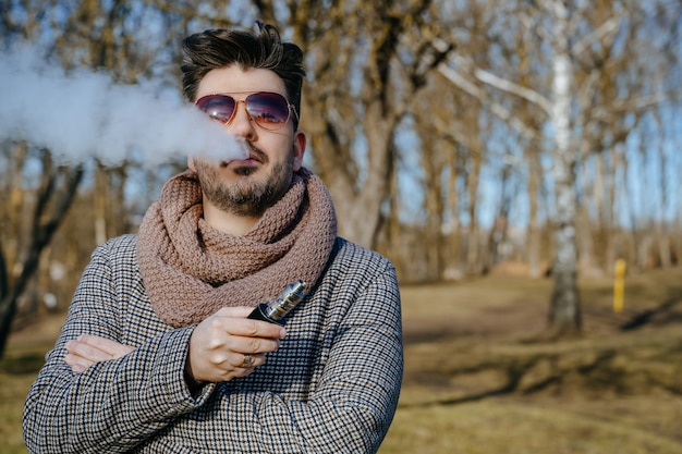 Homem barbudo jovem confiante em cigarro eletrônico de vape de óculos de sol. Um homem jovem bonito barbudo hipster vaping cigarro ao ar livre no parque. Fechar-se.