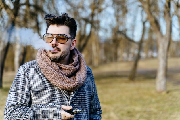 Homem barbudo jovem confiante em cigarro eletrônico de vape de óculos de sol. Um homem jovem bonito barbudo hipster vaping cigarro ao ar livre no parque. Fechar-se.