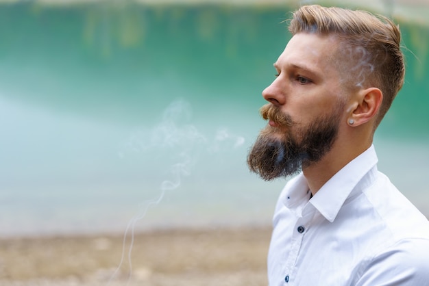 Homem barbudo jovem de negócios caucasiano em uma camisa branca fuma um cigarro e sopra a fumaça contra o ...