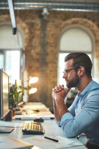 Homem barbudo jovem e bem-sucedido com óculos e roupa formal olhando para o monitor do computador