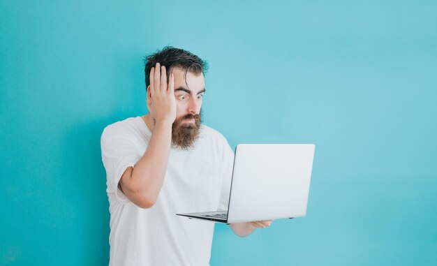 Foto homem barbudo jovem hippie surpreso ao ver o laptop, espaço de cópia, fundo azul suave removível, básico mínimo, estudante, conceito de empreendedor, espaço de camisa branca