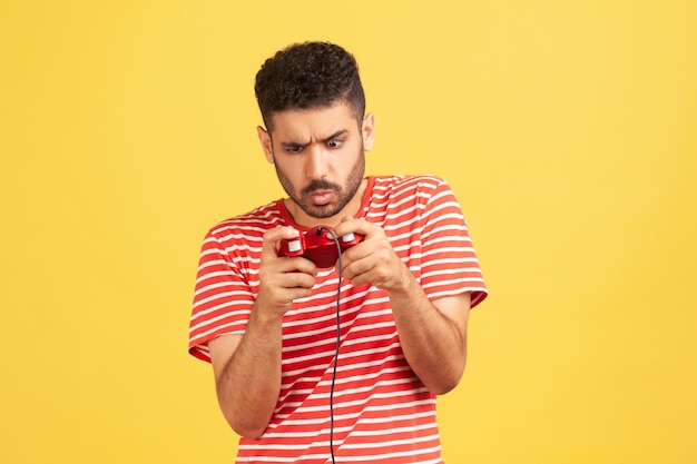 Homem barbudo nervoso viciado em camiseta listrada vermelha fazendo careta segurando joystick, jogando videogame online, competição virtual. Tiro de estúdio interior isolado em fundo amarelo