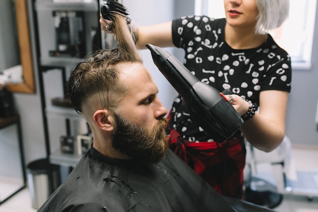 Homem barbudo no cabeleireiro com secador de cabelo enquanto está sentado na cadeira na barbearia.