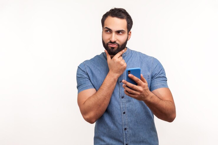 Foto homem barbudo pensativo, coçando o queixo, segurando o smartphone na mão, contemplando a atualização do software, escolhendo as tarifas adequadas, tiro de estúdio interno isolado no fundo branco