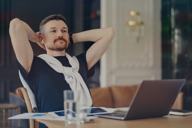 Foto homem barbudo pensativo pondera sobre a tarefa, mantém as mãos atrás da cabeça, senta-se na mesa com aparelhos modernos pensa sobre a distância trabalho concentrado na distância usa camiseta com suéter amarrado sobre os ombros