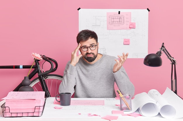 Homem barbudo perplexo levanta a mão mantém a mão nas faces do templo tarefa difícil não consegue encontrar solução usa poses de roupas casuais em um espaço de coworking contra a parede rosa. Arquiteto homem hesitante.