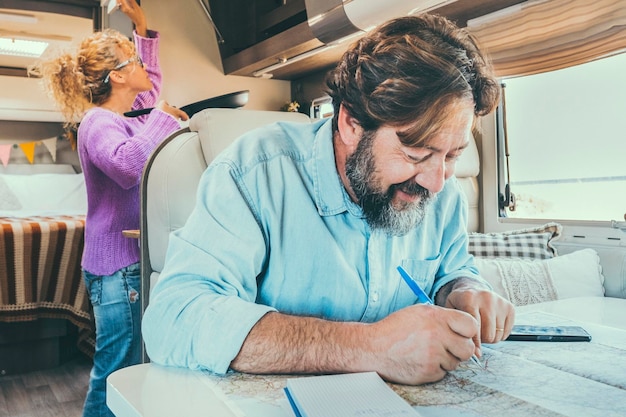 Homem barbudo planejando o próximo destino viagem férias férias sentado à mesa dentro de sua caravana Mulher em segundo plano cozinhando e gerenciando armazenamento Férias férias de vida
