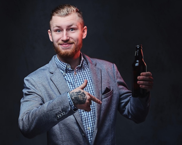 Homem barbudo ruiva elegante vestido de terno detém uma garrafa com cerveja artesanal sobre fundo cinza.