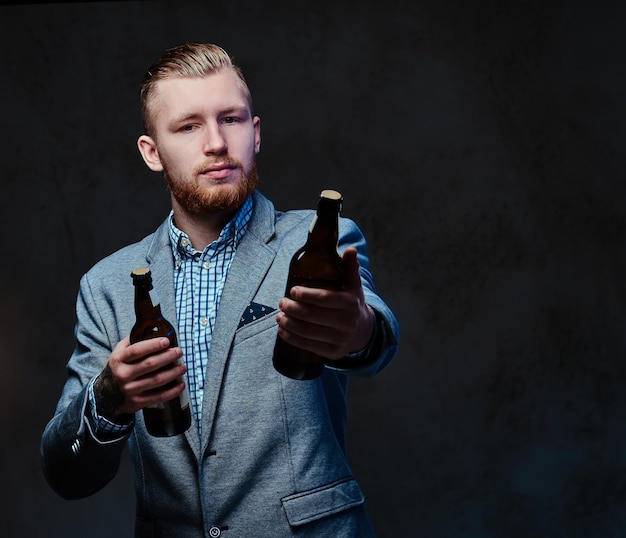 Homem barbudo ruiva elegante vestido de terno detém uma garrafa com cerveja artesanal sobre fundo cinza.