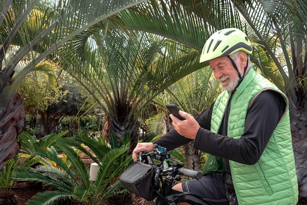 Homem barbudo sênior ativo com capacete andando de bicicleta em um parque tropical olhando para o celular