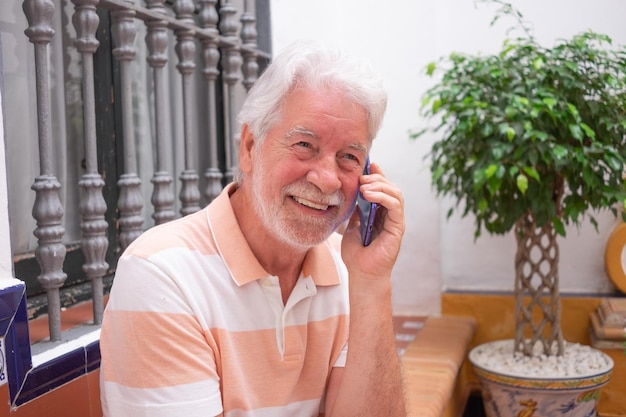 Homem barbudo sênior atraente falando no celular sentado no pátio de casa sorrindo aposentado idoso desfrutando de tecnologia e social