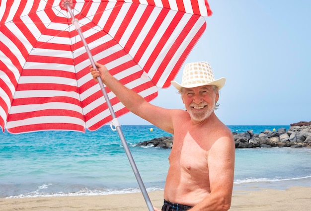 Homem barbudo sênior atraente usando um chapéu de palha curtindo férias na praia segurando um maiô guarda-chuva listrado e grande sorriso ativo aposentado idoso e conceito divertido