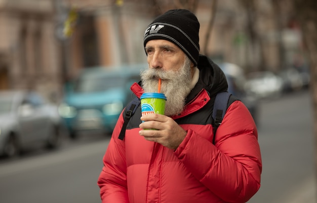 Homem barbudo sênior bonito com copo de papel de café da manhã andando na rua.