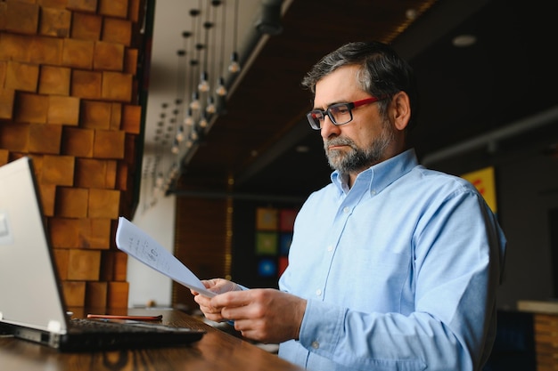 Homem barbudo sênior positivo com cabelos grisalhos, bebendo café e usando laptop no espaço de cópia de café Elegante empresário envelhecido na jaqueta bordô, desfrutando de seu chá enquanto trabalhava on-line