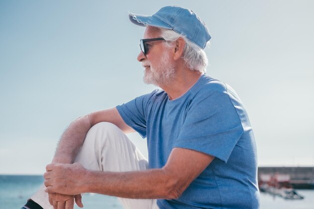Homem barbudo sênior sorridente sentado ao ar livre de frente para o mar desfrutando de férias de viagem de dia ensolarado