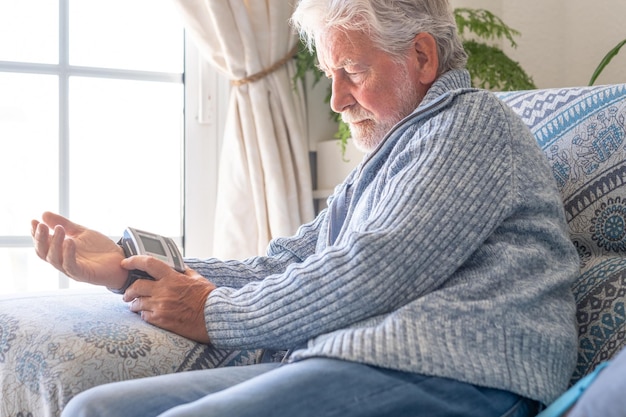 Homem barbudo sênior usando dispositivo médico para medir a pressão arterial avô mais velho sentado em casa no sofá