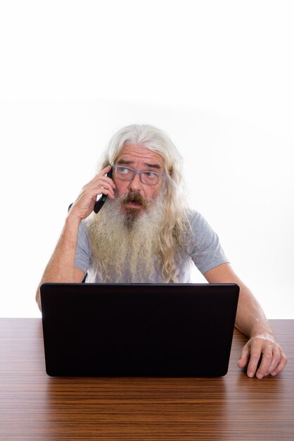 homem barbudo sênior usando óculos e falando no telefone enquanto usa o laptop