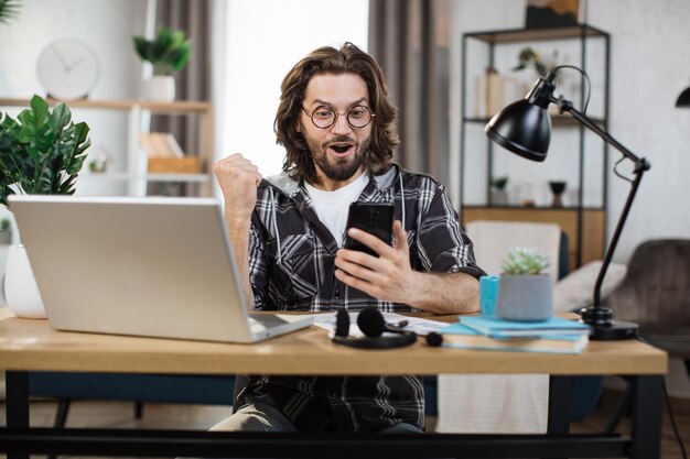 Homem barbudo sentado na mesa e recebe boas notícias trabalhando em smartphone