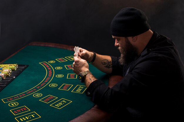 Homem barbudo sério sentado à mesa de pôquer e segurando cartas isoladas em preto. Copie o espaço