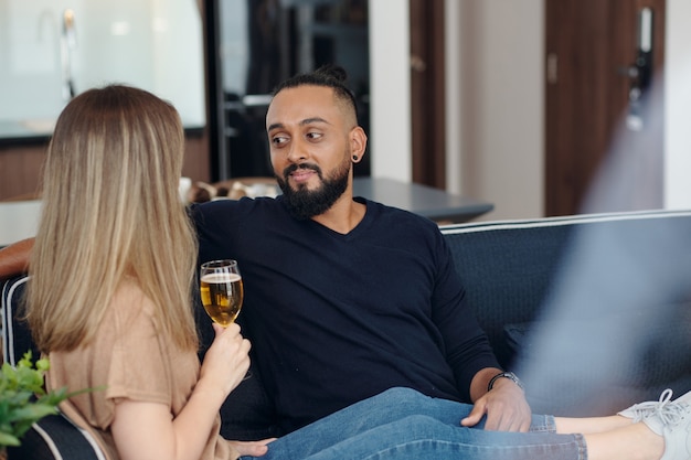 Homem barbudo sorridente descansando no sofá com a namorada bebendo champanhe e contando sobre o dia dela