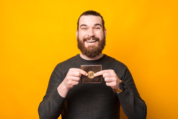 Homem barbudo sorridente em pé sobre uma parede amarela e mostrando bitcoin perto da carteira