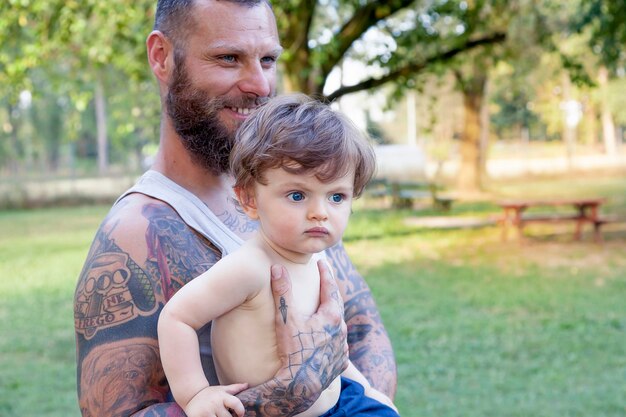 Foto homem barbudo sorridente segurando filho de pé no parque