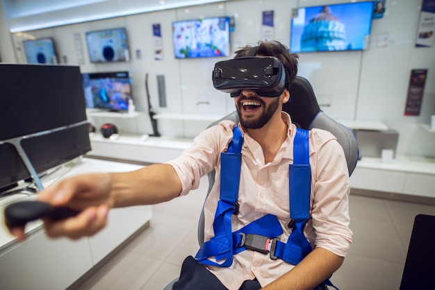 homem barbudo sorridente segurando o joystick sem fio na mão enquanto testa óculos de Vr em uma loja de tecnologia.