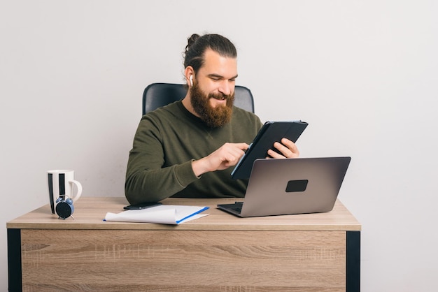 Homem barbudo sorridente, trabalhando no escritório está usando seu tablet ao abrir o laptop.