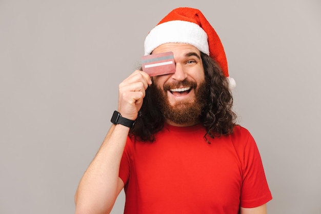 Homem barbudo sorridente usando boné de natal cobre um olho com um cartão vermelho