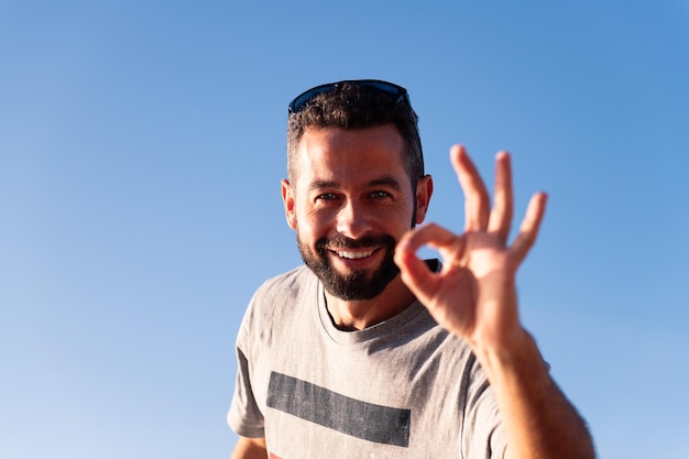 Homem barbudo sorrindo olhando para a câmera fazendo o símbolo OK com a mão com o céu azul no espaço de cópia de fundo para texto