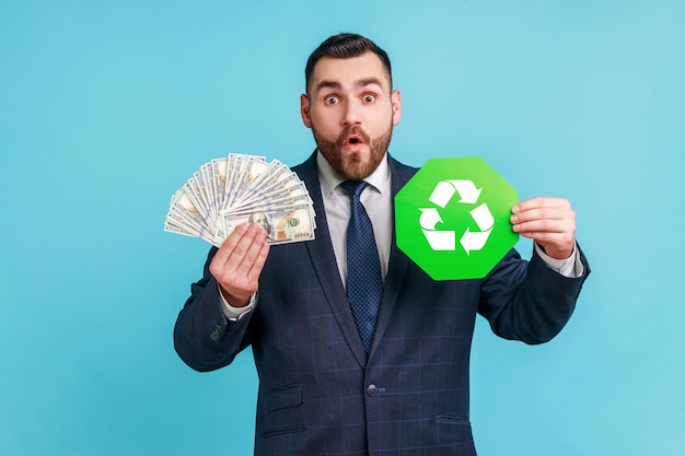 Foto homem barbudo surpreso vestindo terno preto estilo oficial segurando notas de dólares e reciclagem verde canta nas mãos, emoção de expressão. tiro de estúdio interior isolado sobre fundo azul.