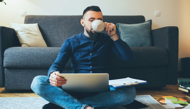Homem barbudo trabalhando em casa no chão com um computador e documentos enquanto bebe um café