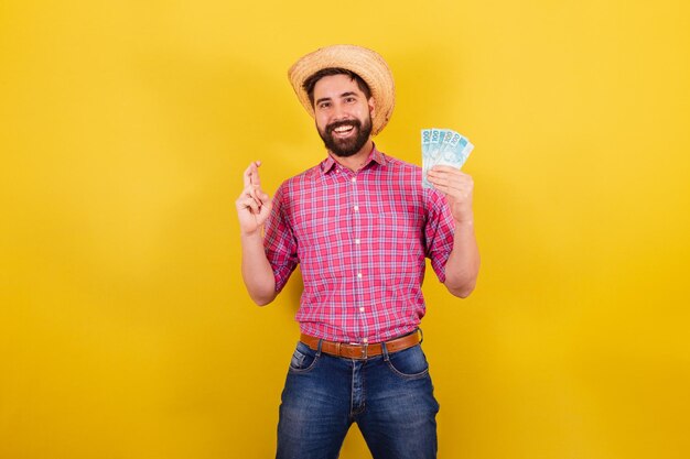 Homem barbudo vestindo roupas típicas para festa Junina cruzando os dedos torcendo e desejando segurando dinheiro dinheiro notas sorte para a festa Arraia