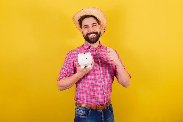 Homem barbudo vestindo roupas típicas para festa Junina segurando moeda e cofrinho finanças e economia para o Festival do Arraia