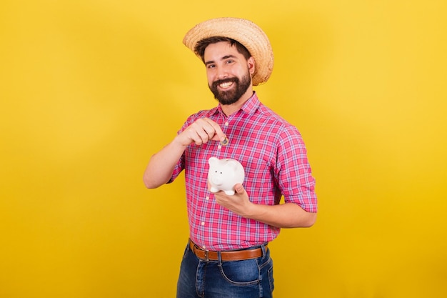 Homem barbudo vestindo roupas típicas para festa Junina segurando moeda e cofrinho finanças e economia para o Festival do Arraia