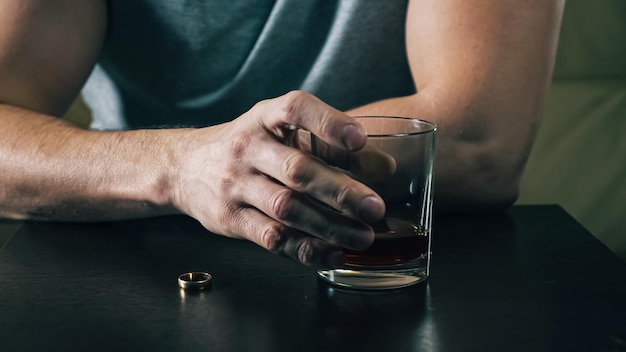 Homem bêbado vestindo camiseta cinza casual olha para o anel de casamento depois de brigar hesitando e decide se divorciar de copo de uísque closeup